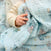 A baby's hand rests on an aden + anais essentials cotton muslin swaddle blanket in light blue with cartoon animals and stars, while wearing a beige long-sleeve, creating a cozy and breathable scene. Available in a 4 pack.