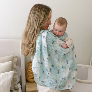 A woman with long blonde hair holds a baby wrapped in an aden + anais essentials cotton muslin swaddle blanket, standing in a cozy, softly lit room with neutral decor.