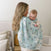 A woman with long blonde hair holds a baby wrapped in an aden + anais essentials cotton muslin swaddle blanket, standing in a cozy, softly lit room with neutral decor.