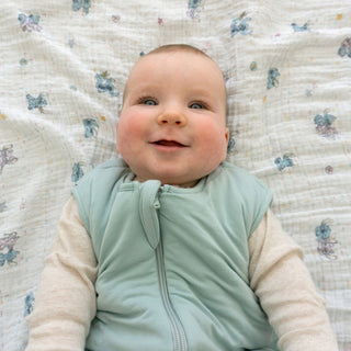 A smiling baby with blue eyes and rosy cheeks lies on the aden + anais essentials cotton muslin swaddle blanket, featuring a light butterfly pattern. The soft, breathable 4-pack makes naptime cozy and comfortable.