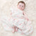 A baby, with rosy cheeks and light brown hair, rests contently on a fluffy white carpet, wrapped in an aden + anais essentials cotton muslin swaddle blanket from their 4 pack, featuring a soft floral pattern.