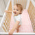 A curly-haired, blue-eyed toddler stands cheerfully in a wooden crib, wearing a white onesie. The crib features an aden + anais essentials cotton muslin crib sheet and a pink patterned mattress.