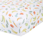 A close-up of the white aden + anais essentials cotton muslin crib sheet reveals a playful print with colorful fruits and vegetables like bananas, carrots, peas, and apples, adding a bright and whimsical touch to the bedding.
