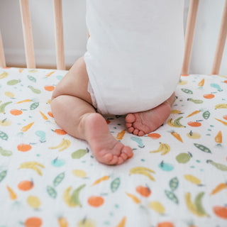A baby in a white onesie kneels on an aden + anais essentials cotton muslin crib sheet, featuring vibrant fruits and vegetables like bananas, carrots, peas, and oranges. The breathable fabric ensures comfort while the wooden bars of the crib can be seen behind.