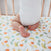 A baby in a white onesie kneels on an aden + anais essentials cotton muslin crib sheet, featuring vibrant fruits and vegetables like bananas, carrots, peas, and oranges. The breathable fabric ensures comfort while the wooden bars of the crib can be seen behind.