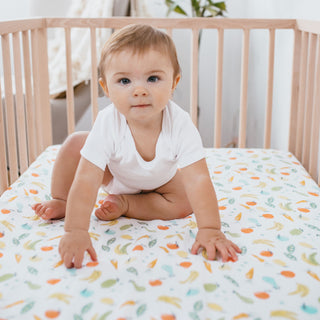 A light brown-haired baby sits in a wooden crib on an aden + anais essentials cotton muslin crib sheet featuring vegetable illustrations. The baby wears a white onesie and gazes curiously at the camera in a softly lit, uncluttered nursery.