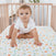 A light brown-haired baby sits in a wooden crib on an aden + anais essentials cotton muslin crib sheet featuring vegetable illustrations. The baby wears a white onesie and gazes curiously at the camera in a softly lit, uncluttered nursery.