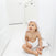 A baby sits on a white floor, wrapped in an aden + anais essentials cotton muslin hooded towel, in a bright white room. The absorbent fabric keeps the little one cozy as they look upward with curiosity.