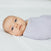 A baby wrapped in a light purple aden + anais essentials cotton muslin swaddle blanket from the 4-pack lies on a white surface, gazing to the side with wide eyes. The infants head is slightly turned, and soft lighting creates a calm atmosphere.