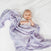 On a white bed, a baby is wrapped in an aden + anais essentials cotton muslin swaddle blanket with a leaf design. The light purple muslin, made from soft cotton, complements the white onesie as the baby smiles with hands raised near their chest.
