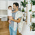 In a modern kitchen with white cabinetry and elegant wood flooring, a man in jeans and a gray T-shirt holds his baby. Draped over his shoulder is a colorful essentials cotton muslin cloth from the aden + anais 4 pack, while vibrant greenery adorns the walls vertical plant holder.