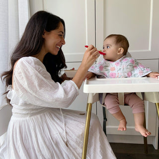 A woman in a white dress smiles while feeding a baby with a red spoon. The baby, wearing an aden + anais boutique 100% cotton muslin floral bib from a 2-pack, sits in a high chair. They are in a bright room with white cabinets and curtains.
