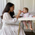 A woman in a white dress smiles while feeding a baby with a red spoon. The baby, wearing an aden + anais boutique 100% cotton muslin floral bib from a 2-pack, sits in a high chair. They are in a bright room with white cabinets and curtains.