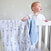A baby in a white outfit stands in a crib, gazing curiously. The aden + anais boutique cotton muslin dream blanket, extra soft and adorned with blue and white dinosaurs, drapes over the rail. Light-colored walls with subtle star patterns enhance the cozy atmosphere.