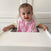 A baby sits in a high chair wearing an adorable aden + anais pink 100% cotton muslin snap bib with adjustable fit and white patterns, adding charm as the baby smiles at the camera. A white cabinet is in the background.