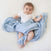 A baby in a white onesie lies on a white surface, wrapped in an aden + anais boutique 100% cotton muslin swaddle blanket with light blue and white stars, looking upward with a slight smile and short, dark hair.