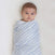 A baby lies on a soft white surface, wrapped in a breathable aden + anais boutique muslin swaddle blanket with light blue and white stripes, looking directly at the camera.