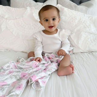 A baby in a white outfit sits on a white bed, holding an aden + anais boutique 100% cotton muslin swaddle blanket adorned with pink patterns, smiling and looking forward.