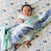 A baby in a white onesie lies on a patterned elephant blanket, partially covered by an aden + anais boutique silky soft muslin dream blanket, featuring bamboo-infused viscose with blue and green swirls, smiling and looking upward.