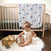 A baby sits on a silky soft, fuzzy white rug, playing with wooden toys. Behind them is a light wooden crib with an aden + anais boutique silky soft muslin dream blanket featuring elephants draped over the side. The room includes a bamboo-infused wooden chair and hardwood floor.