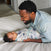 A man in a light denim shirt smiles at a baby swaddled in an aden + anais boutique silky soft swaddle blanket, lying on a white bed. The bamboo-infused viscose blanket makes them both look comfortable and content.