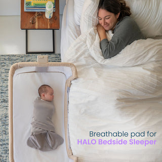 A swaddled infant sleeps on a Bedside Sleeper DreamWeave Breathable Pad in a bedside sleeper next to a parent in bed.