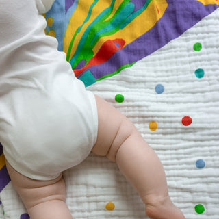 A baby’s legs and torso are visible as they lie on the aden + anais essentials cotton muslin dream blanket, featuring polka dots and abstract patterns.