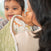 A woman cradles a baby wrapped in an aden + anais Messy Babe Bundle, featuring playful giraffes. The baby beams with joy, nestled indoors in the cozy softness of their special blanket.
