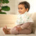 A toddler with short brown hair sits on a beige couch, wearing a pink ribbed outfit and an aden + anais essentials cotton muslin burpy bib with floral design. The child looks to the side with a slight smile, while a green potted plant is visible in the background.