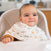 A baby sits in a high chair, wrapped in the Messy Babe Bundle by aden + anais. The child gazes slightly off-camera with a gentle smile, while the blurred background enhances the cozy indoor ambiance.