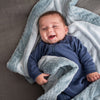 A baby in a soft, blue outfit lies peacefully on a textured gray blanket, smiling beneath an aden + anais essentials plush blanket. The modern colors add gentle charm to the soft gray background.