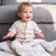 A baby with light hair sits on an essentials plush blanket by aden + anais in front of a gray sofa, wearing a buttoned beige outfit. The cheerful baby looks up as sunlight softly illuminates the cozy scene.