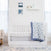 A charming nursery with a white crib featuring a blue and white patterned quilt from the aden + anais essentials cotton muslin bedding set 3 pack. The room includes a light rug, armchair, wall decor with two framed prints, and a shelf displaying an elephant figurine.