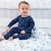 A toddler in dark blue long-sleeve pajamas sits on an animal-patterned blanket. In the nursery background, a crib features the aden + anais essentials cotton muslin bedding set with blue stars. The child smiles and looks slightly to the side.