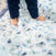 A child stands on the essentials cotton muslin bedding set by aden + anais, featuring animal illustrations in soothing blue and green tones, ideal for the nursery. The cozy quilt showcases delicate muslin details as their dark blue pants reveal bare feet.