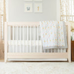 A cozy room features a light-colored crib with white bars adorned by the aden + anais essentials cotton muslin bedding set. It stands on a soft white carpet, with three cute animal prints above and soft curtains framing the windows, creating a serene space.