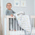 A toddler stands in a crib, holding a colorful muslin blanket. The nurserys light walls feature framed animal prints of an elephant and giraffe above. An aden + anais essentials cotton muslin bedding set 3 pack adds coziness to the charming space.