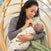 A woman with long dark hair sits on a wooden chair, tenderly holding a swaddled baby in an aden + anais essentials cotton muslin swaddle blanket. She gazes lovingly at the baby, brushing their chin softly as sunlight streams through the window, enveloping them in warmth.