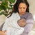 A woman in a gray outfit cradles a sleeping baby wrapped in an aden + anais essentials cotton muslin swaddle blanket featuring patterns. She gazes affectionately at the baby, with a green plant visible in the background.