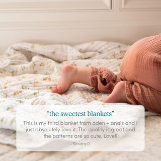A baby in rust ribbed pants lies on an aden + anais essentials cotton muslin dream blanket, featuring a delicate white floral pattern. A customer review overlay praises the blanket’s quality and beautiful design.