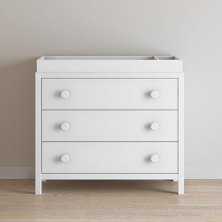 A matte white, three-drawer dresser stands on a light wood floor by a beige wall, topped with the Child Craft F01792 Universal Changing Table Topper.