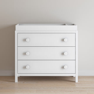 A matte white, three-drawer dresser stands on a light wood floor by a beige wall, topped with the Child Craft F01792 Universal Changing Table Topper.