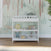 The Child Craft Flat Top Baby Changing Table in matte white, featuring three shelves with baby-safe finishes, provides nursery storage beneath a pastel safari animal mural with giraffes, ostrich, and cheetah. Sunlight shines on the wooden floor.