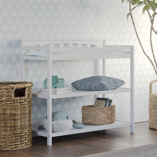 The Child Craft Flat Top Baby Changing Table in matte white features baby-safe finishes and storage shelves for baskets, books, toys, and cushions. It stands beside a woven hamper in a softly lit nursery with patterned wallpaper and a potted plant.