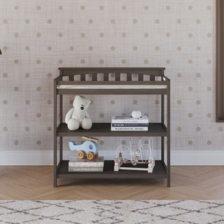 The Child Craft Flat Top Baby Changing Table in dapper gray features a baby changing pad, shelves with a teddy bear, books, a blue elephant toy, and bead maze, set against a pink polka dot wall and patterned rug on wooden flooring.