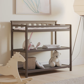 The Child Craft Flat Top Baby Changing Table in Dapper Gray features a baby changing pad and two shelves for nursery storage displaying a plush rabbit, blankets, books, and a teddy bear. A wooden rocking horse and floor lamp complete the softly lit room.
