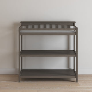 The Child Craft Flat Top Baby Changing Table in Dapper Gray features a cushioned changing pad and two open shelves for nursery storage, shown here against a light wall on a wooden floor.
