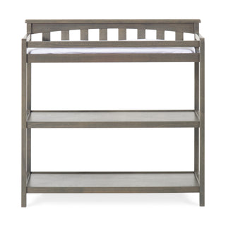 The Child Craft Flat Top Baby Changing Table in Dapper Gray includes a cushioned baby changing pad and two open shelves below for nursery storage, with a flat top design and raised back and sides for safety.