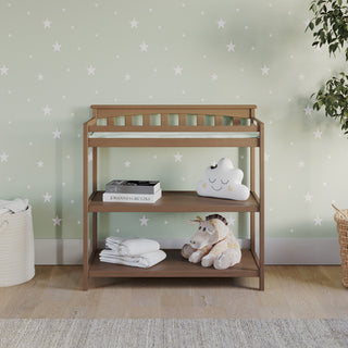 The Child Craft Flat Top Baby Changing Table in Dusty Heather features two shelves for storage, holding a box, cloud pillow, white towels, and plush unicorn. A mint green wall with white stars and a plant on the right add to the cozy nursery feel.