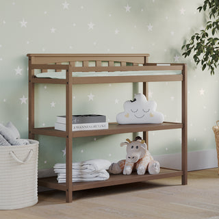 A Child Craft Flat Top Baby Changing Table in Dusty Heather stands against a star wall, topped with a baby changing pad, cloud pillow, books, and giraffe plush. A floor basket with rolled towels adds extra nursery storage nearby.